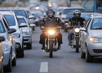 Dois motociclistas de capacete circulam entre carros parados no corredor de trânsito urbano