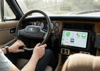 Motorista com mãos no volante de carro vintage com painel de madeira e tela CarPlay mostrando aplicativos de telefone e mensagens