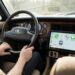 Motorista com mãos no volante de carro vintage com painel de madeira e tela CarPlay mostrando aplicativos de telefone e mensagens