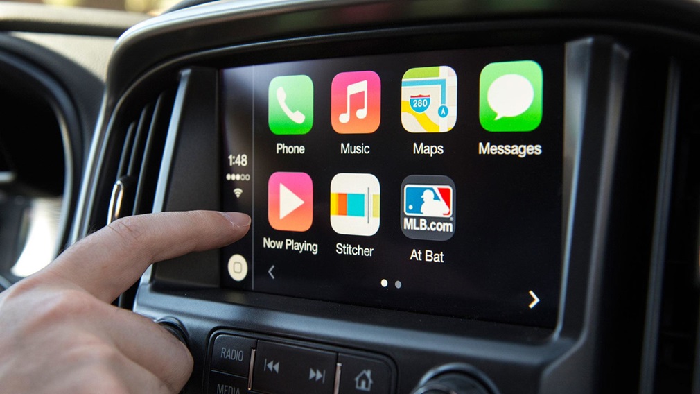 Tela do CarPlay mostrando apps Phone, Music, Maps, Messages, Now Playing, Stitcher e At Bat com dedo tocando