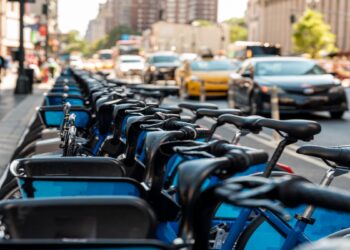 Imagem ilustrativa de fileira de bicicletas compartilhadas estacionadas em estação à beira de avenida movimentada, com carros ao lado, cena usada para tratar das regras de circulação de bicicletas elétricas