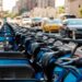 Imagem ilustrativa de fileira de bicicletas compartilhadas estacionadas em estação à beira de avenida movimentada, com carros ao lado, cena usada para tratar das regras de circulação de bicicletas elétricas