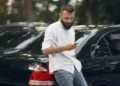 Homem consulta informações no celular ao lado de um carro sobre recall pendente.
