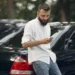 Homem consulta informações no celular ao lado de um carro sobre recall pendente.