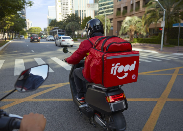 Entregador do iFood circula de moto em avenida na zona urbana.
