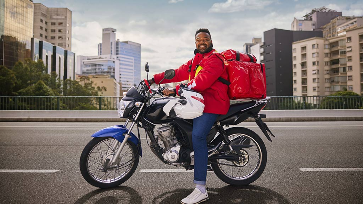 Entregador de aplicativo posa feliz em motocicleta no centro urbano.
