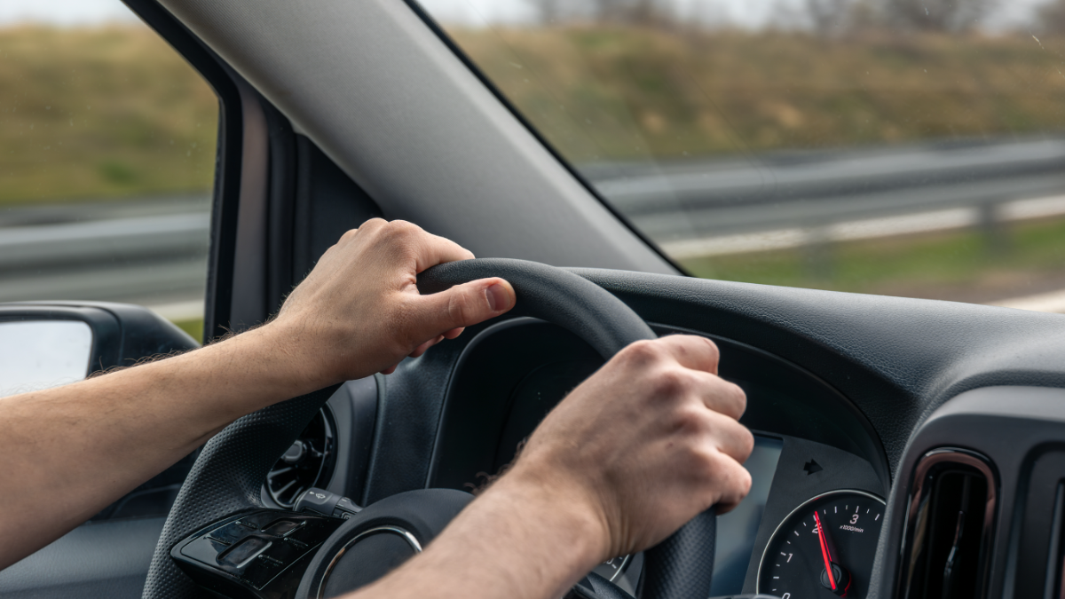 Mãos de adulto, aparentemente homem, segurando volante de veículo em rodovia.