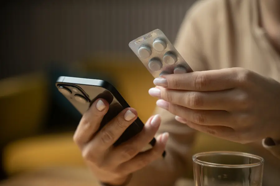 Consumidora consulta celular enquanto segura cartela de medicamentos antes de realizar compra pela internet.