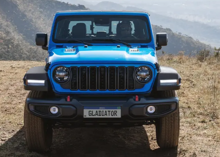 Jeep Gladiator azul visto de frente em terreno de terra com placa personalizada e montanhas ao fundo.