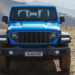 Jeep Gladiator azul visto de frente em terreno de terra com placa personalizada e montanhas ao fundo.