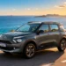 SUV cinza percorre uma estrada litorânea com o mar azul ao fundo, sob a luz intensa do sol.