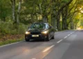 Um carro preto trafega por uma estrada arborizada, com os faróis acesos e a pista cercada por uma vegetação verde e densa.
