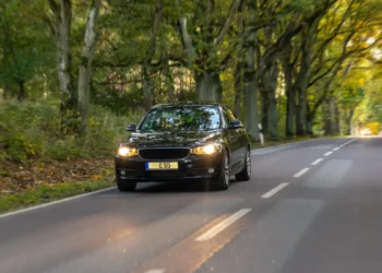 Um carro preto trafega por uma estrada arborizada, com os faróis acesos e a pista cercada por uma vegetação verde e densa.