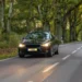 Um carro preto trafega por uma estrada arborizada, com os faróis acesos e a pista cercada por uma vegetação verde e densa.