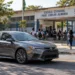 Carro trafega em frente a escola pública com estudantes ao fundo, representando os cuidados com a velocidade em áreas de grande circulação de pessoas.