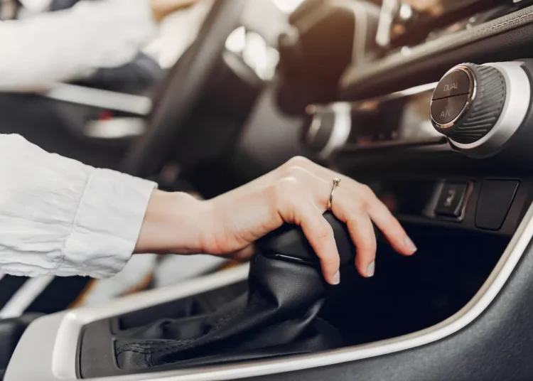 Mão sobre a alavanca de câmbio automático, com o console central do carro ao fundo.