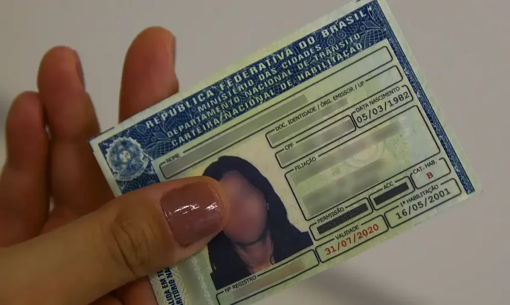 Mão segura uma Carteira Nacional de Habilitação impressa, com a fotografia parcialmente coberta pelo polegar.