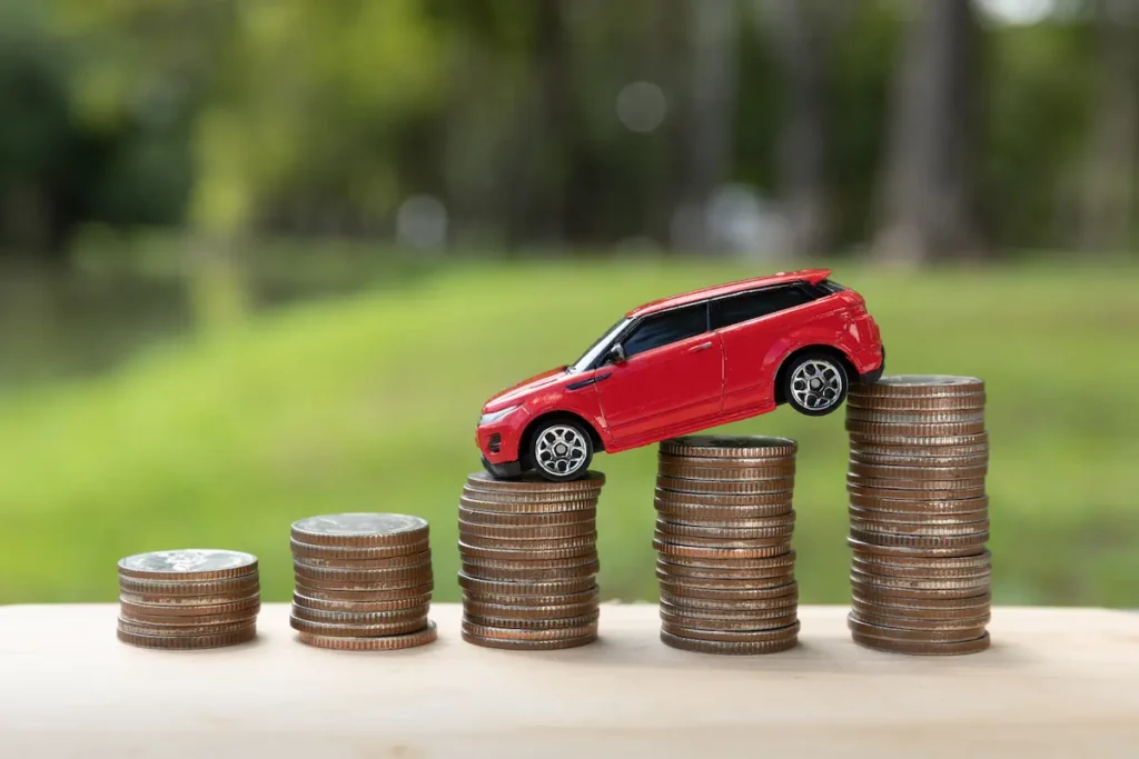 Carro vermelho em miniatura sobre pilhas de moedas de diferentes alturas, simbolizando os custos e a importância do pagamento do licenciamento veicular.