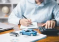 Homem utilizando calculadora e fazendo anotações ao lado de um notebook, uma chave de carro e um veículo em miniatura, representando os custos do licenciamento veicular.