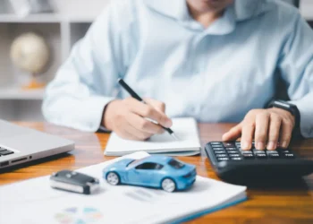 Homem utilizando calculadora e fazendo anotações ao lado de um notebook, uma chave de carro e um veículo em miniatura, representando os custos do licenciamento veicular.