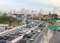 Trânsito intenso em avenida com carros, motos e ônibus circulando entre diferentes faixas.