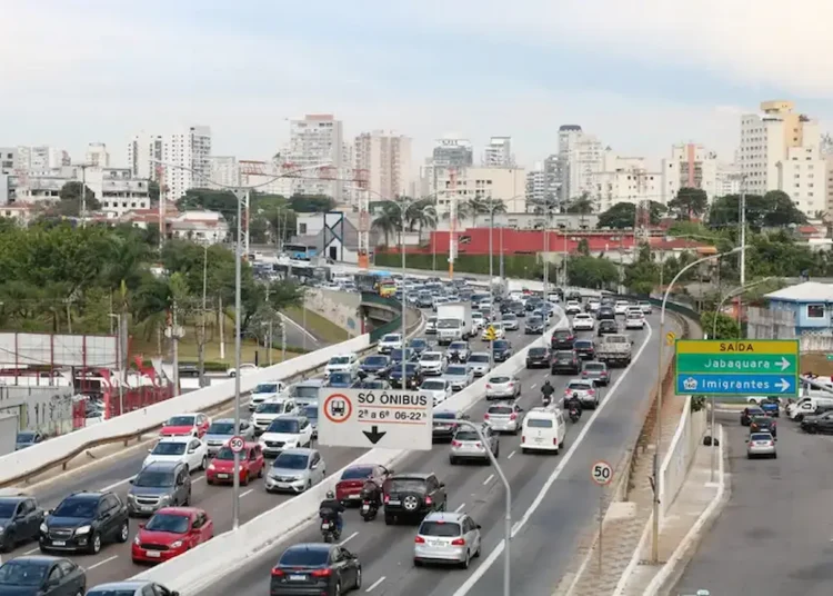 Trânsito intenso em avenida com carros, motos e ônibus circulando entre diferentes faixas.
